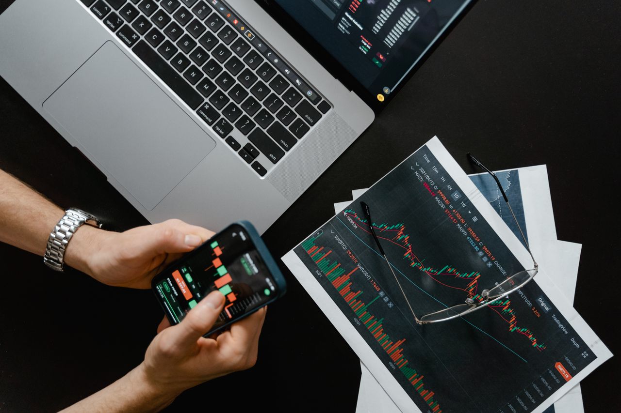 services-02 Des mains utilisant un smartphone à côté d’un ordinateur portable affichant des graphiques boursiers, illustrant le trading numérique.